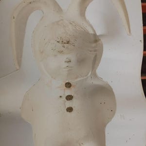 May include: A white plastic mold featuring the recessed shape of a figure wearing a hood with long rabbit ears. The design includes a detailed face and three small circular indentations down the center of the chest area, showing some surface wear.