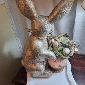 Puede incluir: Una figurita de conejo de pascua de brillo dorado sosteniendo una cesta de huevos de Pascua rosa y dorada llena de acentos florales verdes y blancos.