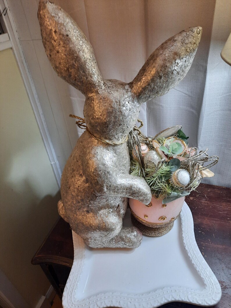 Puede incluir: Una figurita de conejo de pascua de brillo dorado sosteniendo una cesta de huevos de Pascua rosa y dorada llena de acentos florales verdes y blancos.