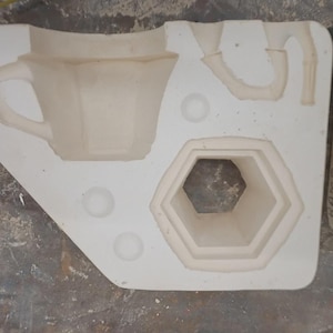 May include: A white plaster ceramic slip casting mold featuring recessed cavities for a hexagonal cup with a handle and a separate handle component. The mold has a flat, rectangular base with rounded corners and three small circular indentations.