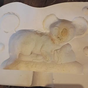 May include: A white plaster mold featuring a detailed relief of a koala bear lying down. The koala is resting on a textured surface, with visible details of its fur and features. The mold is rectangular with rounded edges.