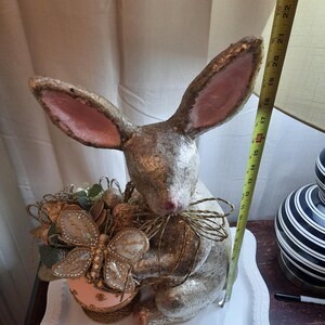 Puede incluir: Una figurita de conejo plateada y rosa que sostiene una mariposa decorativa y un huevo. El conejo est&aacute; sentado en un plato blanco con borde festoneado.