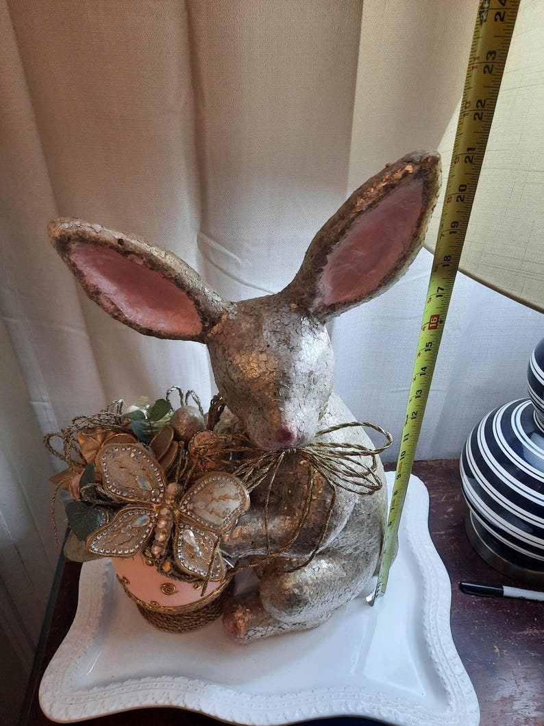 Puede incluir: Una figurita de conejo plateada y rosa que sostiene una mariposa decorativa y un huevo. El conejo est&aacute; sentado en un plato blanco con borde festoneado.