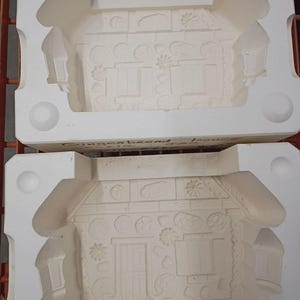 May include: Two white ceramic slip casting molds for a gingerbread house. Each mold features recessed details of a house facade with a door, windows, and decorative elements. The text "Gingerbread house" is written in black ink on the side.
