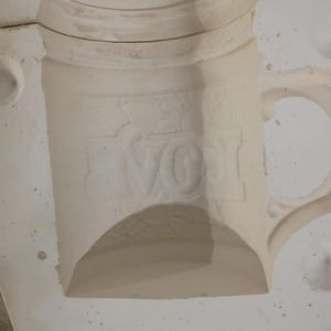 May include: A white ceramic slip casting mold for a mug featuring a debossed design of the word LOVE with a sun icon above the letters. The mold is composed of two interlocking sections with a handle cavity and a cylindrical body shape.