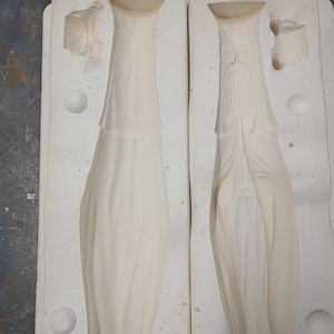 May include: Two off-white ceramic slip casting molds for a tall, slender figure. The molds feature a recessed, elongated design with a flared base and a detailed relief of a bearded face wearing a crown on the upper section of the right mold.