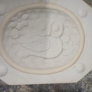 May include: A white ceramic plaster mold featuring an oval recessed area with a relief design of an owl surrounded by flowers. The mold has a textured background and the text MACKY # 124 is clearly printed on the bottom edge of the plaster form.