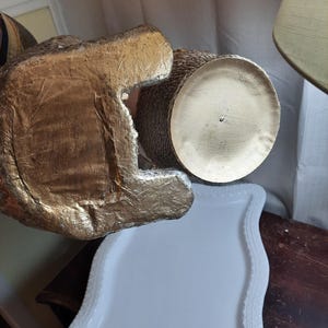 Puede incluir: Un jarr&oacute;n de cer&aacute;mica de color dorado con una superficie texturizada y una base redonda. El jarr&oacute;n est&aacute; sobre una bandeja blanca con un borde decorativo.