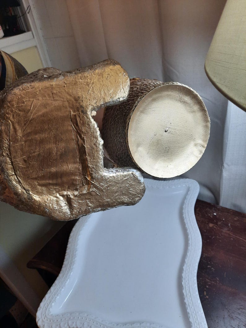 Puede incluir: Un jarr&oacute;n de cer&aacute;mica de color dorado con una superficie texturizada y una base redonda. El jarr&oacute;n est&aacute; sobre una bandeja blanca con un borde decorativo.