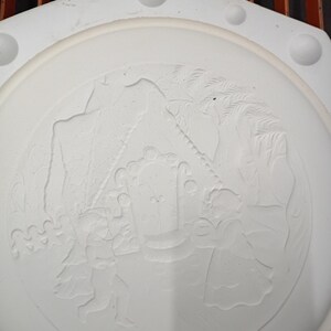 May include: A white, circular ceramic plate with a raised, detailed relief design. The design depicts figures, possibly angels or cherubs, near a structure, possibly a building or tent, with trees and other elements. The plate has a hexagonal outer shape.