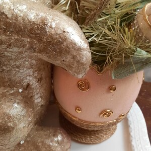 Puede incluir: Una figurita de conejito de brillantina dorada junto a un huevo de Pascua de brillantina rosa con detalles dorados. El huevo est&aacute; sobre una base tejida.