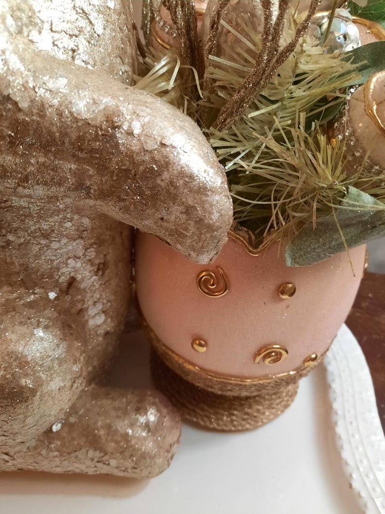 Puede incluir: Una figurita de conejito de brillantina dorada junto a un huevo de Pascua de brillantina rosa con detalles dorados. El huevo est&aacute; sobre una base tejida.