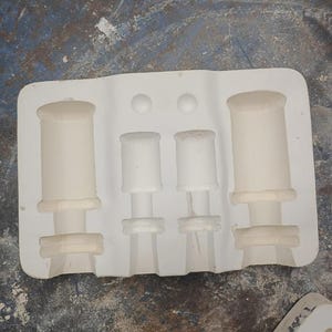 May include: A rectangular white plaster mold featuring four distinct cylindrical cavities of varying sizes. The mold has two larger outer impressions and two smaller inner impressions, each with detailed circular indentations and tiered sections.