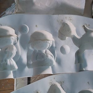 May include: A white plaster mold featuring relief figures of a nativity scene. The mold includes figures of people, a camel, and spherical shapes. The figures are in various stages of completion, with some showing fine details.