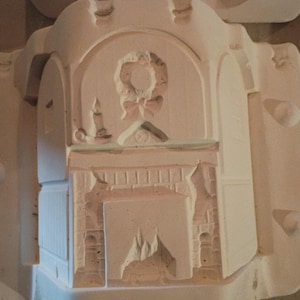 May include: A white plaster mold featuring a fireplace design with a wreath, candle, and arched doorway. The fireplace has a detailed brick pattern and a flame design. The mold is likely for creating decorative items.