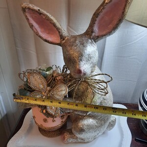 Puede incluir: Una figurita de conejo de papel mach&eacute; plateado y rosa que sostiene un huevo de Pascua rosa y dorado con una decoraci&oacute;n de mariposa. El conejo est&aacute; sentado en un plato de cer&aacute;mica blanco.