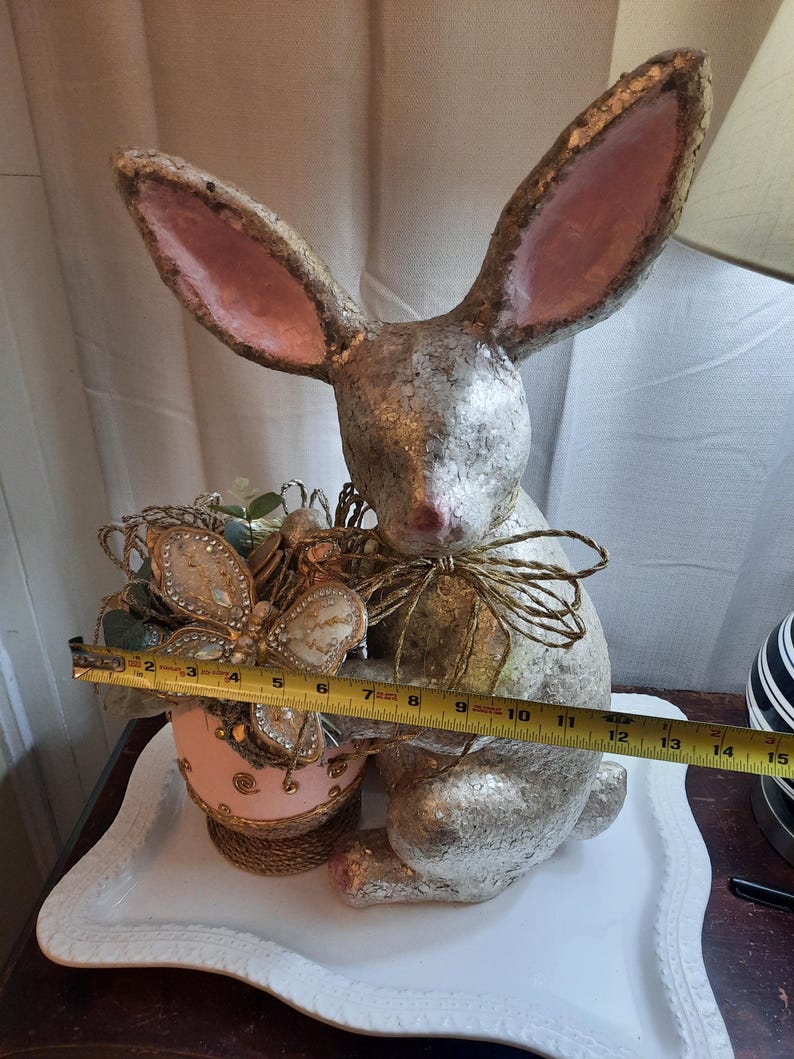 Puede incluir: Una figurita de conejo de papel mach&eacute; plateado y rosa que sostiene un huevo de Pascua rosa y dorado con una decoraci&oacute;n de mariposa. El conejo est&aacute; sentado en un plato de cer&aacute;mica blanco.
