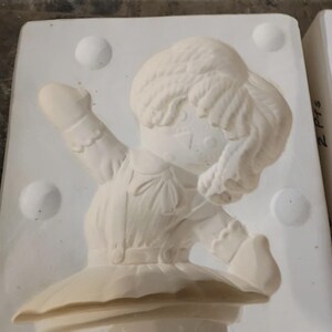 May include: A white ceramic slip mold featuring a recessed design of a doll with yarn-style hair, a collared shirt, and a skirt. The mold has a rectangular base with four circular indentations and the text 2 Pts written on the side edge.
