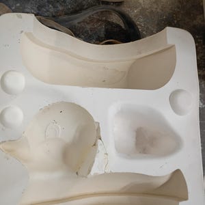 May include: A white plaster ceramic slip casting mold featuring a recessed cavity shaped like a duck. The mold has a smooth, matte surface with circular registration points on the sides and a distinct impression of a duck's head and body.