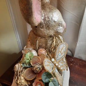 Puede incluir: Una figurita de conejo dorada y blanca con detalles de mariposa y flores. El conejo est&aacute; sentado sobre una superficie blanca con una superficie de madera marr&oacute;n en el fondo.