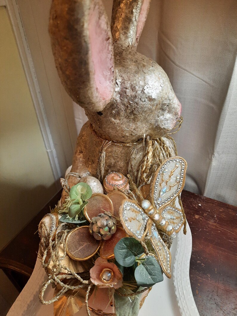 Puede incluir: Una figurita de conejo dorada y blanca con detalles de mariposa y flores. El conejo est&aacute; sentado sobre una superficie blanca con una superficie de madera marr&oacute;n en el fondo.