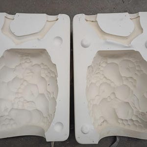 May include: Two white plaster molds, each with a detailed, textured interior. The molds are open, revealing the negative space of a rounded, organic form. The top section of each mold has a curved, antler-like shape.