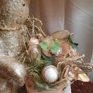 Puede incluir: Una figurita de conejo de Pascua dorada y rosa con un nido de paja, vegetaci&oacute;n y huevos decorativos. El conejo sostiene un huevo rosa y dorado con un borde dorado.