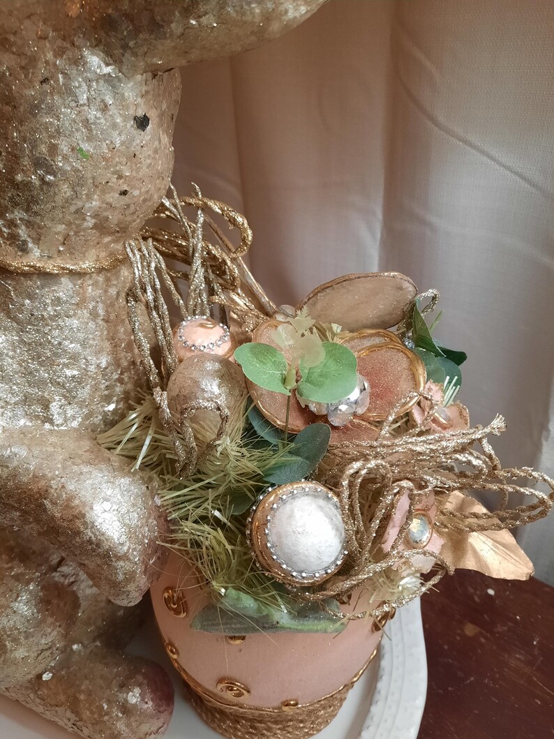 Puede incluir: Una figurita de conejo de Pascua dorada y rosa con un nido de paja, vegetaci&oacute;n y huevos decorativos. El conejo sostiene un huevo rosa y dorado con un borde dorado.