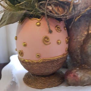 Puede incluir: Un huevo de Pascua rosa con detalles dorados y una base tejida. El huevo est&aacute; decorado con espirales y puntos dorados.