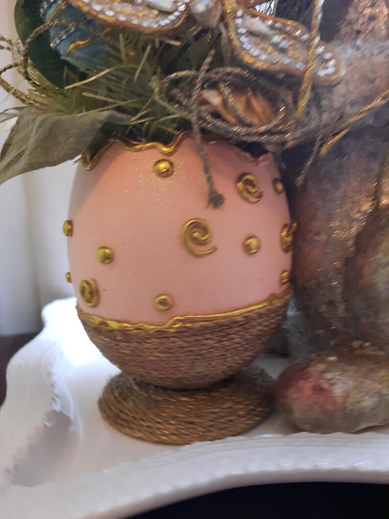 Puede incluir: Un huevo de Pascua rosa con detalles dorados y una base tejida. El huevo est&aacute; decorado con espirales y puntos dorados.