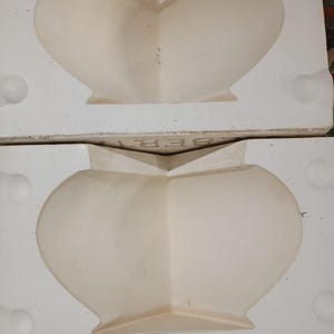 May include: A two-part white ceramic slip casting mold for a vase or vessel. The interior features a smooth, curved cavity with a wide opening. The exterior block has circular registration indentations and the partial text BER printed on the top edge.