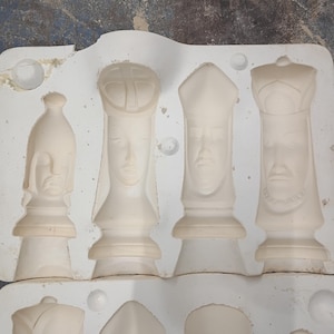 May include: A white chess piece mold with detailed facial features. The mold contains several chess pieces, including a king, queen, bishop, and rook. The pieces are in various stages of completion, showing the intricate design of the chess set.