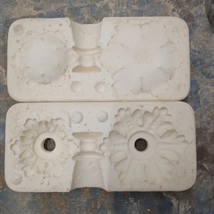 May include: Two rectangular off-white plaster molds featuring intricate floral and leaf-like relief patterns. The molds contain recessed circular cavities and detailed impressions designed for casting decorative elements with central holes.