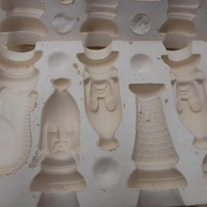 May include: A white ceramic slip casting mold featuring recessed cavities for creating chess pieces. The mold includes negative impressions of a knight, a bishop, a rook, and other game piece shapes with detailed textures and facial features.