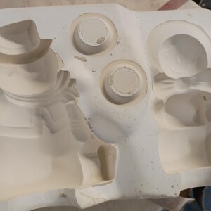 May include: A white ceramic slip casting mold featuring recessed cavities for a snowman figure wearing a hat and scarf, a separate figure with a bow, and two small circular shapes. The mold has a smooth, matte finish and defined edges.