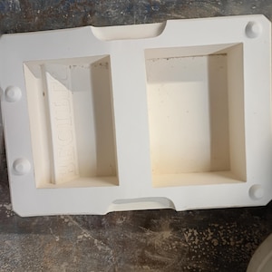 May include: A rectangular white plaster mold featuring two hollowed-out compartments. The left compartment contains the embossed text "BEGIBEE" in reverse. The mold has four circular registration points at the corners and indented side grips.