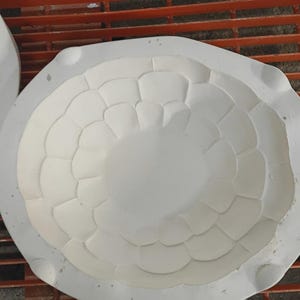 May include: A white plaster ceramic mold featuring a circular, concave interior with a repeating, layered petal or scale pattern radiating from the center. The mold has a smooth, matte finish and a thick, rounded outer rim with small indentations.