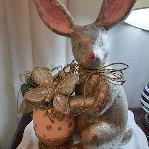 Puede incluir: Una figurita de conejo de pascua de brillo dorado con orejas rosas sosteniendo un huevo de Pascua rosa y dorado con una decoraci&oacute;n de mariposa dorada.