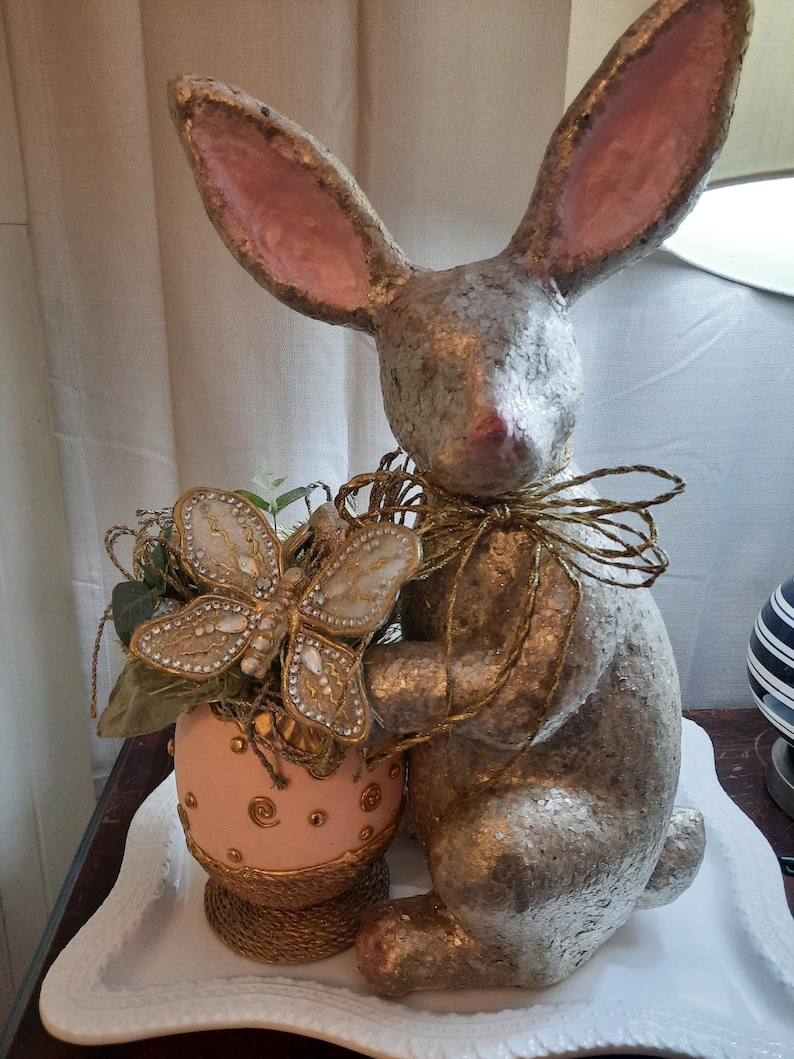 Puede incluir: Una figurita de conejo de pascua de brillo dorado con orejas rosas sosteniendo un huevo de Pascua rosa y dorado con una decoraci&oacute;n de mariposa dorada.