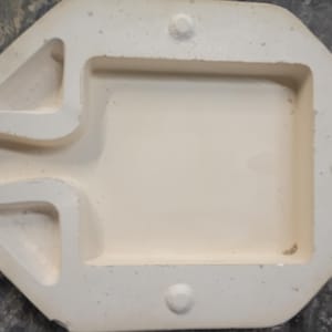 May include: A cream-colored ceramic slip casting mold featuring a central rectangular cavity with a handle-like extension on one side. The mold has a smooth, matte finish with three circular registration points along the outer edges for alignment.