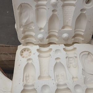 May include: A white plaster mold for casting chess pieces, featuring recessed cavities for a knight, a rook, a bishop, and other game components. The mold is split into two sections with detailed negative impressions of the various chess figures.