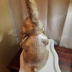 Puede incluir: Una figurita de conejo dorada y blanca con un acento floral verde. El conejo est&aacute; sentado en un pedestal blanco con un borde decorativo.