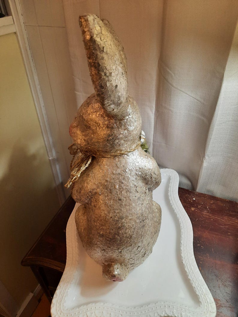 Puede incluir: Una figurita de conejo dorada y blanca con un acento floral verde. El conejo est&aacute; sentado en un pedestal blanco con un borde decorativo.