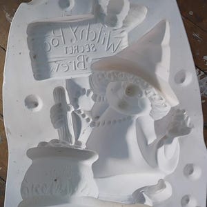 May include: A white plaster mold featuring a whimsical witch design. The witch wears a pointed hat and holds a cauldron with the text "Hot Cocoa". A sign reads "Witch's Secret Brew". The mold is for crafting decorative items.