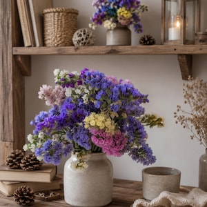May include: A bouquet of dried statice flowers in shades of blue, purple, pink, white, and yellow arranged in a speckled, light-coloured ceramic vase. The flowers feature small, papery petals and are displayed on a wooden surface with pinecones.