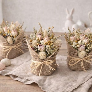 Könnte beinhalten: Drei kleine Blumengestecke in Jutegewickeln, die mit Bindfaden gebunden sind. Jedes Arrangement zeigt getrocknete Rosen, weiße Blüten und Grün. Dekorative Ostereier und ein Keramikkaninchen sind ebenfalls zu sehen.