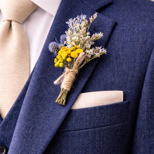 Puede incluir: Un traje azul marino con corbata y pañuelo de bolsillo beige. Un pequeño ramo de flores amarillas, blancas y azules está prendido en la solapa. Las flores están atadas con hilo, creando un aspecto rústico y elegante.