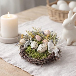 May include: An Easter still life arrangement. It features a white candle, a decorative nest with speckled eggs, a white ceramic bunny, and a basket of white eggs. The items are arranged on a linen tablecloth, creating a soft, inviting atmosphere.