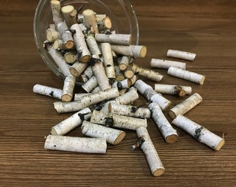 May include: A glass bowl filled with white birch bark pieces spills onto a brown wooden surface. The bark pieces are various lengths and have a natural, rustic look.