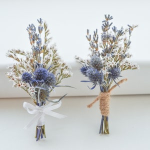 May include: Two small, dried flower bouquets. Each bouquet features blue thistle, lavender, and white star-shaped flowers. One is tied with a white ribbon, the other with twine. Perfect for weddings or special events.